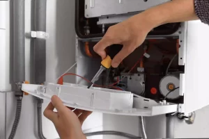 A person uses a screwdriver on a wall-mounted boiler, adjusting components inside