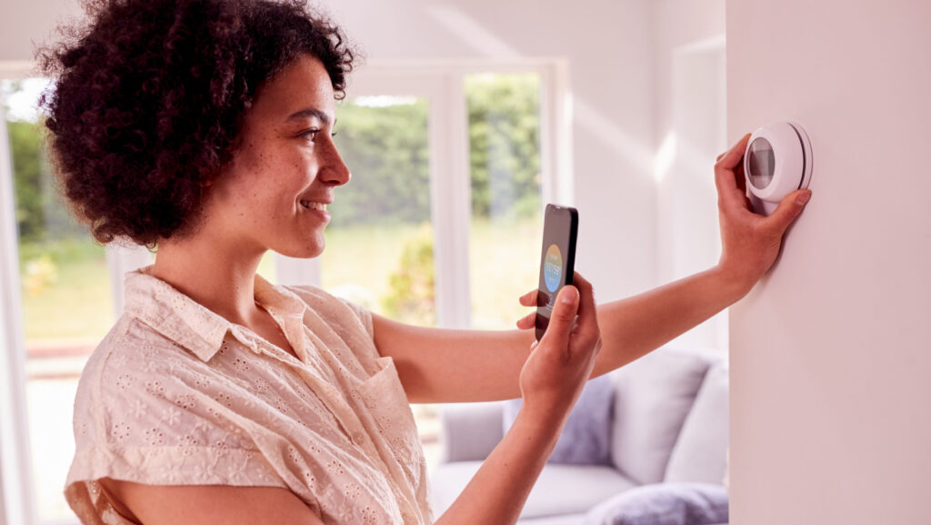 Woman with curly hair adjusting thermostat while holding a phone.